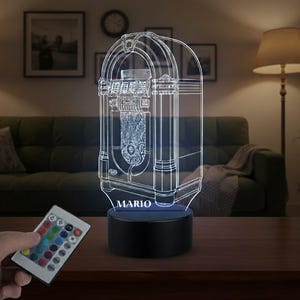 Peut inclure: Lampe LED 3D en forme de juke-box vintage, avec le nom "MARIO" à la base. La lampe émet une lumière douce et brillante. Une télécommande est visible, suggérant la possibilité de personnaliser la couleur.