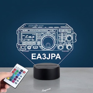 Pode incluir: Uma lâmpada com um desenho a traço branco de um transceptor Yaesu HF/VHF/UHF FT-897, iluminada por uma base preta. O texto "EA3JPA" está abaixo. Um controle remoto com várias opções de cores está em primeiro plano.