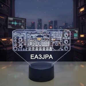 Peut inclure: Lampe en forme de transceiver radio de style néon avec une base noire. La lampe présente un contour détaillé du panneau avant de la radio, comprenant des boutons, des cadrans et un écran. Le texte "EA3JPA" est affiché en dessous.