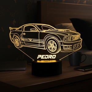 Könnte beinhalten: Ein nachtlicht in Form eines Autos mit 3D-Effekt, das ein warmes gelbes Leuchten ausstrahlt. Der Name "PEDRO" ist an der Basis beleuchtet. Das Design ist eine detaillierte Umrisszeichnung eines Sportwagens.