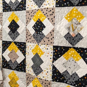 Puede incluir: Una colcha con un patrón geométrico en tonos de gris, negro, blanco y amarillo. La colcha está compuesta por cuadrados con un patrón de diamante dentro de cada cuadrado. La colcha está colgada en una pared de ladrillo blanco.