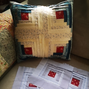 May include: Two blue and white striped fabric squares with red polka dot squares in the centre, and a larger blue and white striped fabric square with red polka dot squares in the centre. The squares are arranged on a beige sofa with a cushion behind them. The cushion is made of patchwork fabric with a log cabin pattern in blue, white, and red.