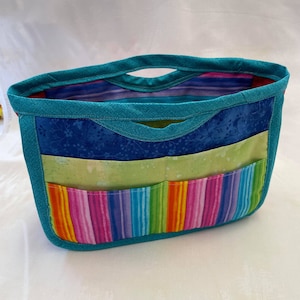 Puede incluir: Un organizador de tela azul turquesa con múltiples bolsillos. Los bolsillos están forrados con una variedad de telas coloridas, incluyendo una tela a rayas verde y blanca, una tela floral azul y blanca y una tela a rayas arcoíris.