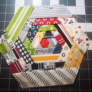 Puede incluir: Un bloque de colcha de retazos en forma de hexágono con una variedad de telas coloridas. Las telas presentan diferentes patrones, incluyendo estrellas, rayas y diseños a cuadros. El bloque está formado por 12 piezas individuales de tela, cada una con un patrón diferente. El bloque está cosido en espiral, con el centro de la espiral siendo una pequeña pieza de tela blanca. El bloque está rodeado por un borde blanco.