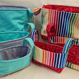 May include: Four fabric storage baskets with handles. The largest basket is striped in rainbow colors. The other three baskets are smaller and have a gray and teal polka dot pattern.