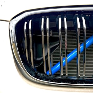 Könnte beinhalten: Nahaufnahme des Kühlergrills eines Autos. Der Kühlergrill hat vertikale Chromstangen vor einem schwarzen Hintergrund. Hinter dem Kühlergrill ist ein hellblauer Balken zu sehen. Die Karosserie des Autos ist weiß.