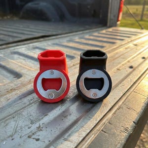 May include: Two bottle openers, one red and one black, with silver bottle opener components. The openers are made of a rubbery material and are sitting on a metal surface. The background is a truck bed.