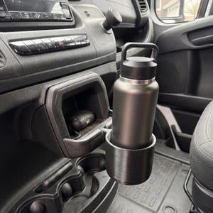 May include: A black insulated water bottle with a black handle sits in a custom-fit cup holder inside a vehicle. The cup holder is mounted on the dashboard, next to a storage compartment. A car key is visible in the compartment.