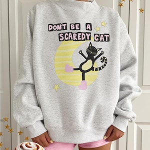 May include: A light gray heathered sweatshirt featuring a graphic of a black cat wearing pink boots, positioned in front of a yellow crescent moon. The text "DON'T BE A SCAREDY CAT" is printed above the cat in black letters with pink outlines.