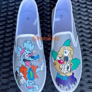 Könnte beinhalten: Ein Paar graue Slip-on-Canvas-Schuhe mit handbemalten Comicfiguren. Ein Schuh zeigt eine blau-grüne zombieartige Kreatur mit einem roten Shirt und einer grünen Hose. Der andere Schuh zeigt eine gelbe Comicfigur mit grünen Haaren und einer blauen Fliege. Die Schuhe sind Größe 9.