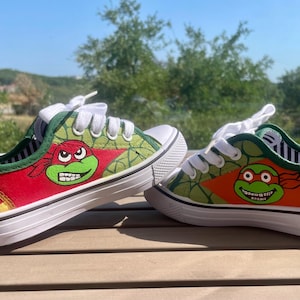 Könnte beinhalten: Ein Paar handbemalte Leinenschuhe mit Designs der Teenage Mutant Ninja Turtles. Die Schuhe haben weiße Schnürsenkel und Sohlen mit grünen, roten und orangenen Akzenten. Das Design umfasst Pizzastücke und Schildkrötengesichter.