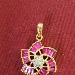 Puede incluir: Pendiente de oro con un diseño de flor rosa y blanca. La flor tiene un centro de pequeñas piedras blancas y está rodeada de piedras rosas.