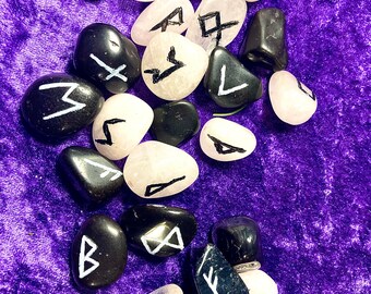Puede incluir: Una colección de piedras negras y blancas con runas pintadas en ellas. Las piedras están esparcidas sobre una superficie de terciopelo morado.