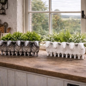 Può includere: Due fioriere decorative a forma di pecora, una argentata e l'altra bianca, riempite di verde, sono appoggiate su un piano di lavoro in legno. Una finestra e utensili da cucina sono sullo sfondo.