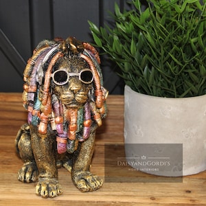 Puede incluir: Estatua decorativa de león dorado y bronce con gafas de sol y rastas coloridas. La estatua tiene detalles y está colocada sobre una superficie de madera. A la derecha hay una planta en maceta con hojas verdes.