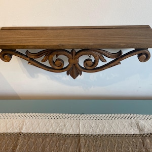 May include: A wooden wall shelf with a decorative, carved scrollwork design. The shelf is a medium brown color and has a flat top. The scrollwork is dark brown and features a central fleur-de-lis motif.