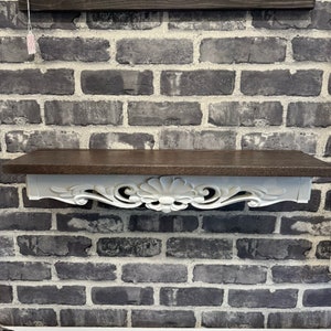 May include: A decorative wall shelf with a dark brown top and a white, ornate floral design on the bottom. The shelf is mounted on a gray brick wall. The shelf is perfect for displaying small items.