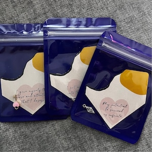 May include: Three blue zippered pouches with white and yellow abstract designs. Each pouch contains a small charm with a different inspirational quote. The first pouch has a pink heart charm with the quote "I give myself the care and attention that I deserve". The second pouch has a silver key charm with the quote "I put my heart into this". The third pouch has a silver heart charm with the quote "My potential to succeed is infinite."