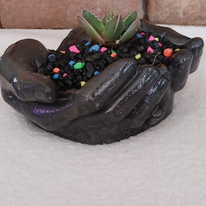 Pode incluir: Vaso de cerâmica preto em forma de duas mãos segurando uma pequena planta suculenta. O vaso está cheio de terra preta e pedras coloridas.