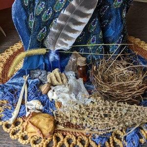 May include: A blue velvet fabric with a beaded pattern, a bird feather, a small bottle, a crystal, a bird's nest, and other natural elements arranged on a woven mat.