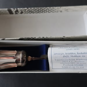 May include: A silver and copper colored metal cup with a handle, in a white box with a blue lining. The box has a label with the text "Van Pallandt sinds 1890 serviezen, bestekken, kookattributen, Delft, Makkum, tin Vijzelstraat/hoek Bakkerstraat 6811 ET Arnhem telefoon 085-42410CT Postbank 831888 NMB 65.42.75.359".