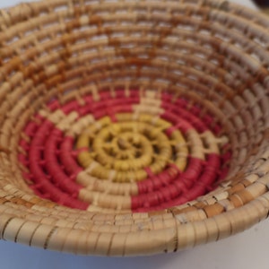 May include: A woven basket with a circular design in the center. The basket is made of natural materials and has a light brown color. The center design features concentric circles in pink and yellow.