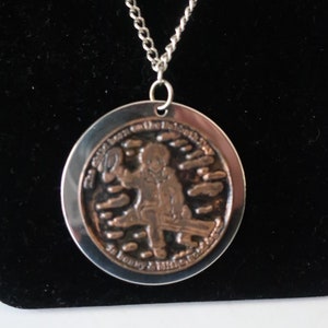 May include: A silver chain necklace with a round pendant featuring a copper-coloured image of a person in a cowboy hat and a quote about the importance of being kind.