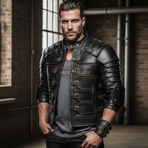 May include: Black leather jacket with multiple buckles and armor-like shoulder detailing. The jacket has a zipper closure and a stand-up collar. The text "MADE HIDES" is visible on the jacket. The model is wearing a gray v-neck shirt and a leather wristband.