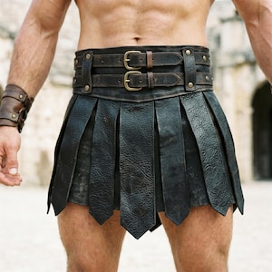 Op de afbeelding: Een zwarte leren gladiatorenrok met een dubbele gespriem. De rok heeft verticale plooien en een decoratieve, met studs versierde riem. Het leer heeft een verweerde uitstraling. De rok is ontworpen om om de taille te worden gedragen.
