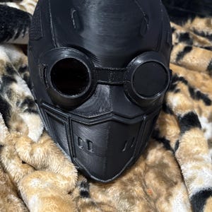 May include: A black, 3D-printed helmet with integrated goggles and a face mask. The helmet features a smooth, matte finish and is designed with a futuristic aesthetic. The goggles have circular lenses, and the face mask has a series of rectangular vents.