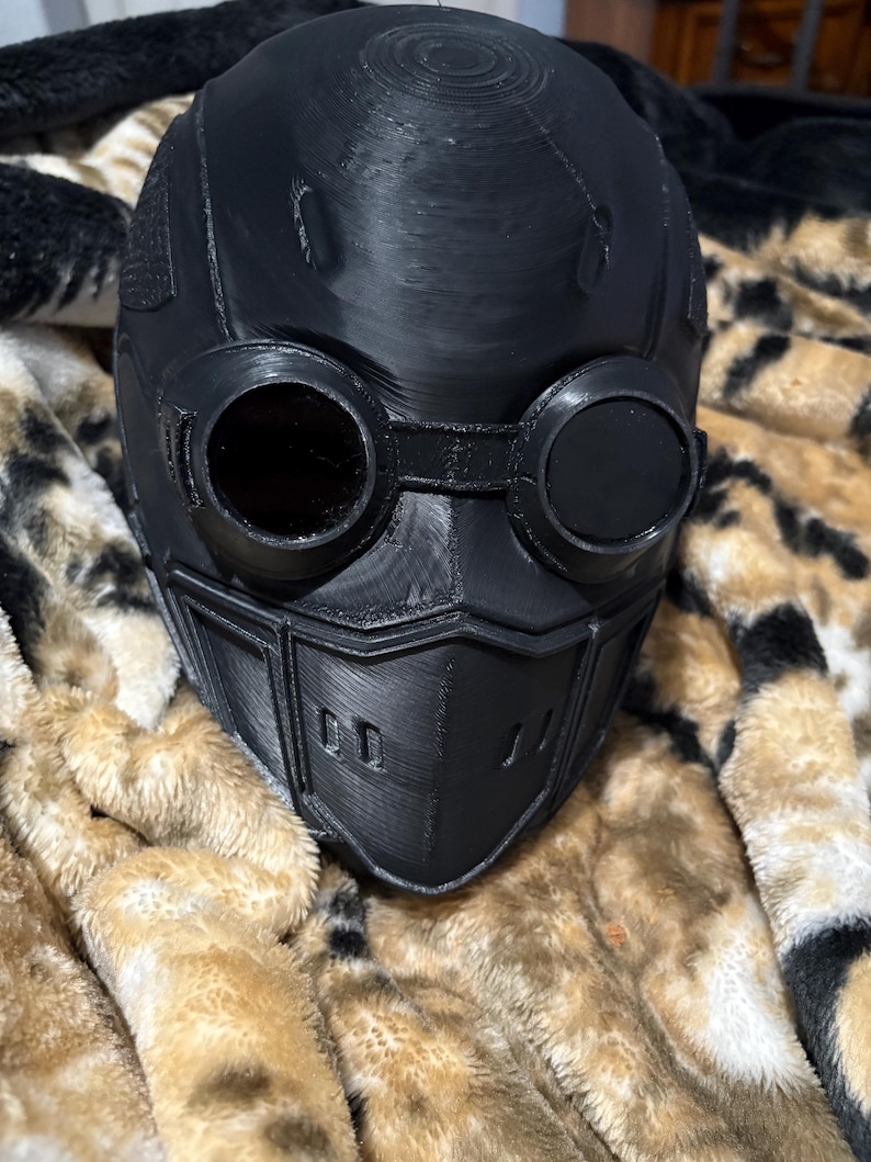May include: A black, 3D-printed helmet with integrated goggles and a face mask. The helmet features a smooth, matte finish and is designed with a futuristic aesthetic. The goggles have circular lenses, and the face mask has a series of rectangular vents.