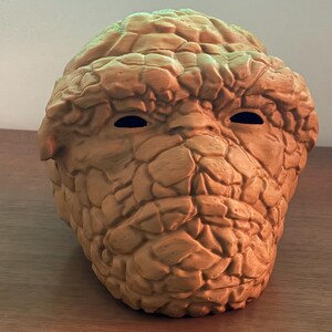May include: A brown, rocky, textured mask of a character with two black eye holes. The mask is made of a hard plastic material.