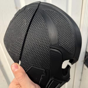 May include: A black, textured mask with a geometric design. The mask is made of a hard material and has a split down the middle. The mask has a smooth, curved shape and is held by a hand.