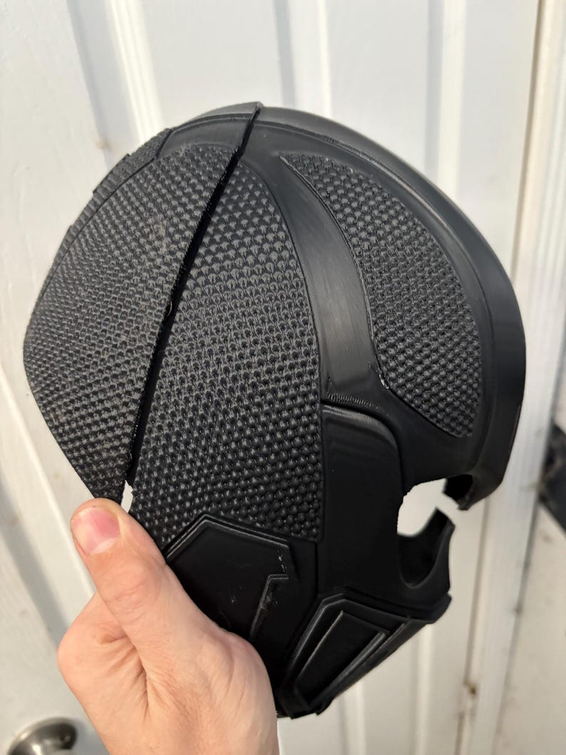 May include: A black, textured mask with a geometric design. The mask is made of a hard material and has a split down the middle. The mask has a smooth, curved shape and is held by a hand.