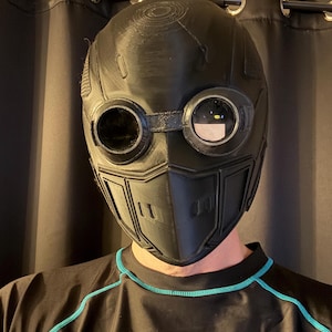 May include: Black, futuristic-style mask with circular goggles and a segmented lower face. The mask has a textured surface and is designed with a series of vents and panels. The goggles have clear lenses. The mask is worn over a black shirt with teal trim.