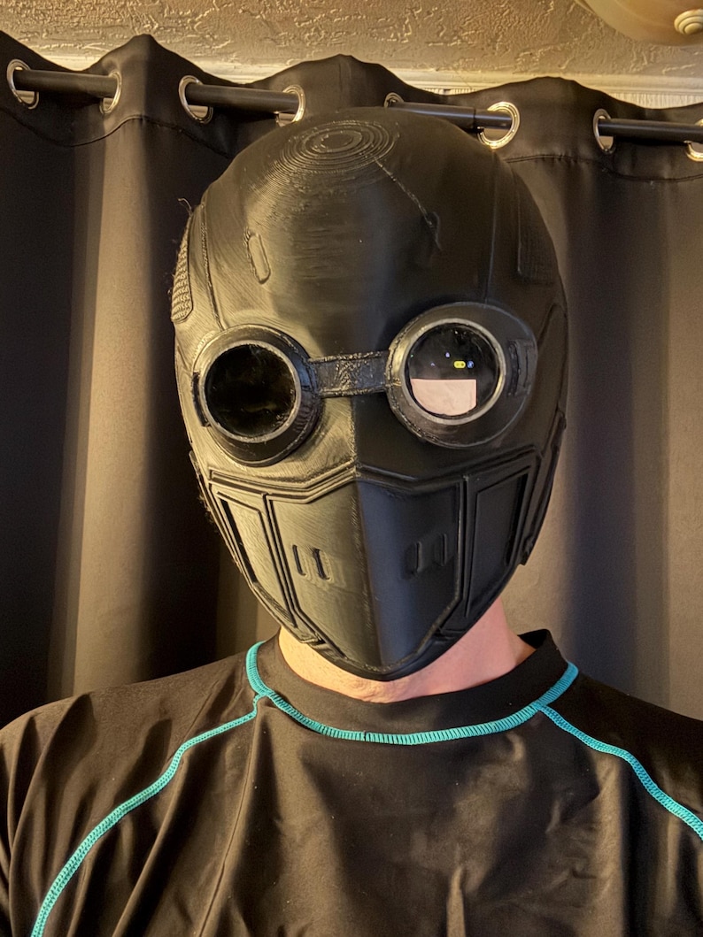 May include: Black, futuristic-style mask with circular goggles and a segmented lower face. The mask has a textured surface and is designed with a series of vents and panels. The goggles have clear lenses. The mask is worn over a black shirt with teal trim.