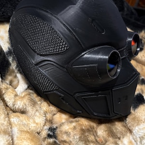 May include: A black, futuristic-looking helmet with integrated goggles. The helmet features textured panels and a smooth, rounded top. The goggles have red lenses. The helmet is resting on a brown and tan patterned blanket.