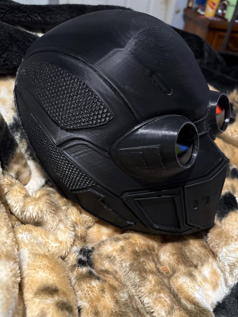 May include: A black, futuristic-looking helmet with integrated goggles. The helmet features textured panels and a smooth, rounded top. The goggles have red lenses. The helmet is resting on a brown and tan patterned blanket.