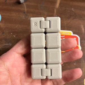 May include: A gray fidget toy with a textured surface and a letter "R" on one side. The toy is made of multiple squares that can be rotated and flipped.