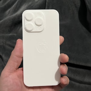 May include: A white iPhone case with a 3D-printed design. The case features a camera module with four circular lenses and an Apple logo. The case is held in a hand against a dark gray background.