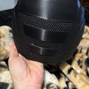 May include: A black helmet with a textured mesh section and smooth, curved panels. The helmet is held in a hand, and the background is a patterned blanket.