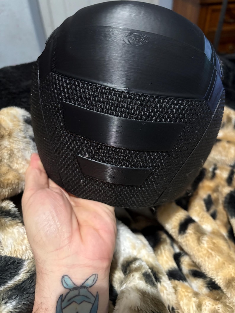 May include: A black helmet with a textured mesh section and smooth, curved panels. The helmet is held in a hand, and the background is a patterned blanket.