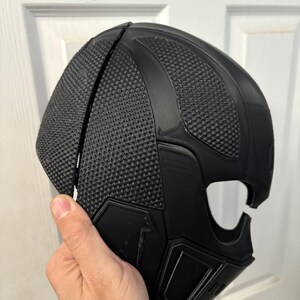 May include: A black, textured mask with a geometric design. The mask has a mesh-like pattern on the top and a smooth, angular design on the lower portion. The mask is held by a hand.