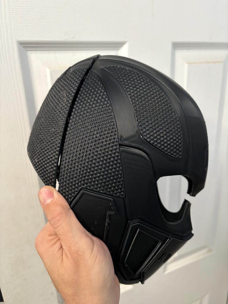 May include: A black, textured mask with a geometric design. The mask has a mesh-like pattern on the top and a smooth, angular design on the lower portion. The mask is held by a hand.