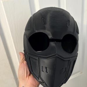May include: Black full-face helmet with large eye openings and a detailed, angular design. The helmet has a textured surface and is likely 3D-printed. The design includes a chin guard with two rectangular vents.