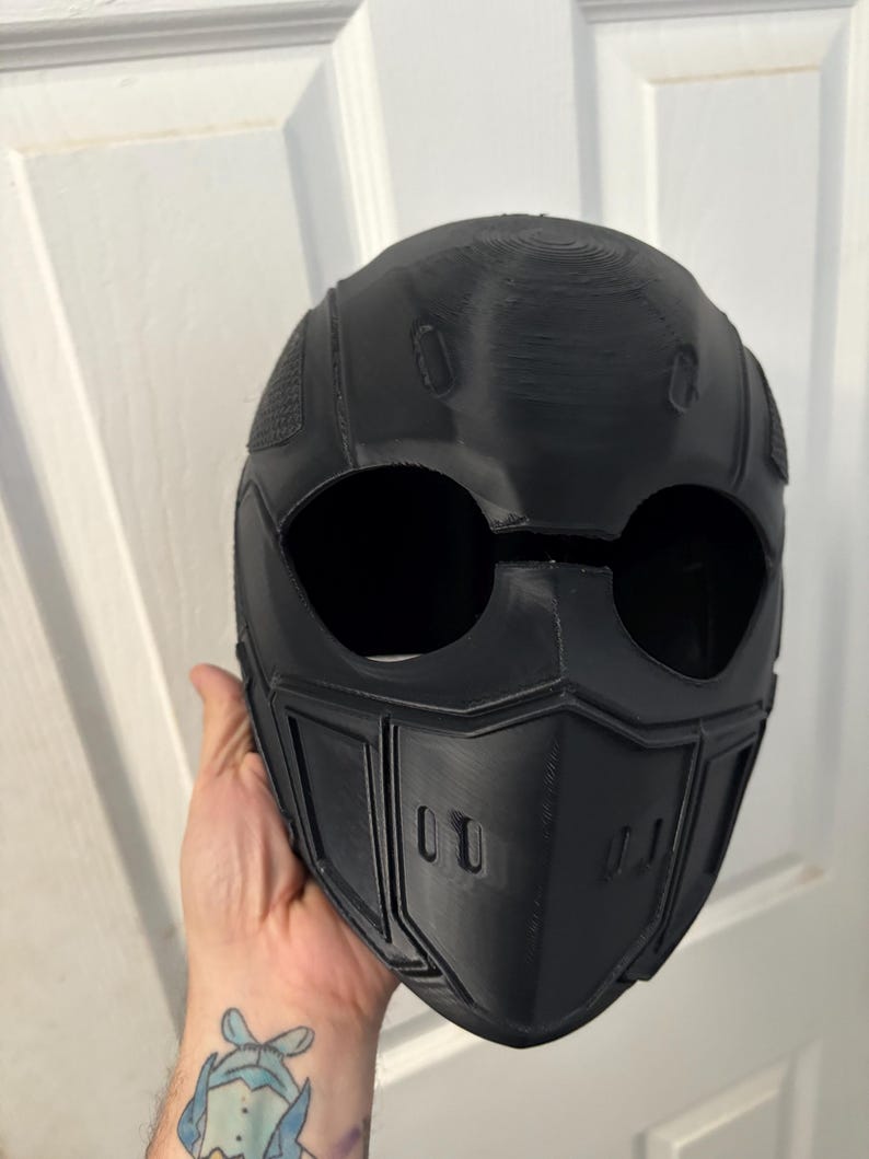 May include: Black full-face helmet with large eye openings and a detailed, angular design. The helmet has a textured surface and is likely 3D-printed. The design includes a chin guard with two rectangular vents.