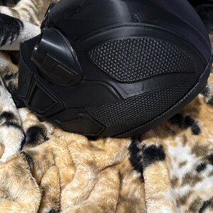 May include: A black, futuristic-looking helmet with textured panels and a visor. The helmet has a smooth, rounded top and angular details around the face and sides. The surface has a matte finish. The helmet is resting on a plush, patterned blanket.
