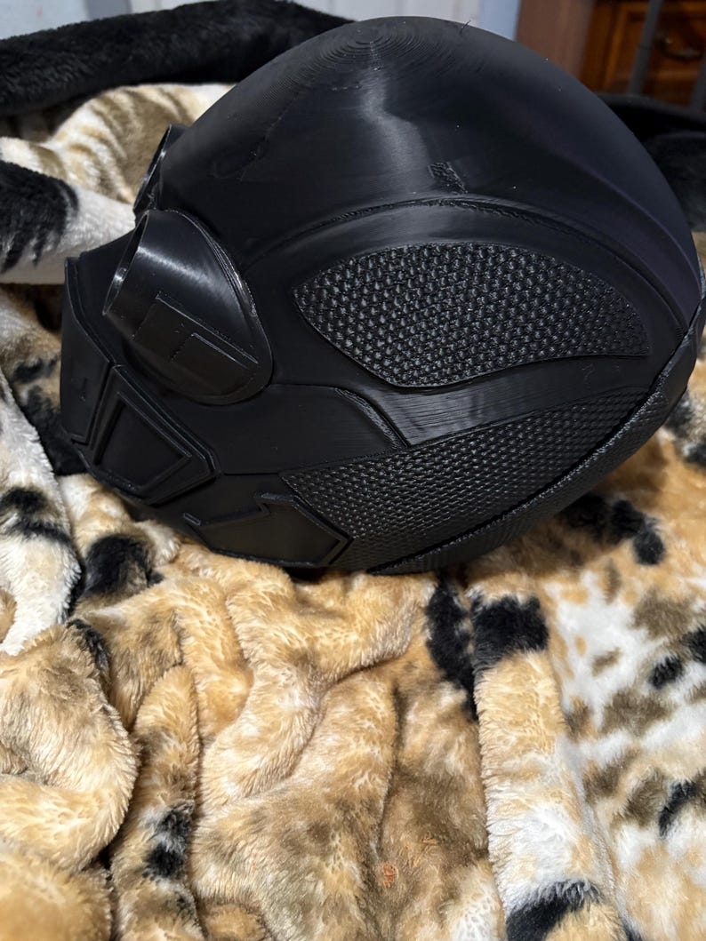 May include: A black, futuristic-looking helmet with textured panels and a visor. The helmet has a smooth, rounded top and angular details around the face and sides. The surface has a matte finish. The helmet is resting on a plush, patterned blanket.