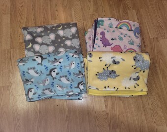 May include: Four fleece blankets with different animal prints. The blankets are folded and stacked on a wooden surface. The prints include elephants, sharks, sheep, and dinosaurs.