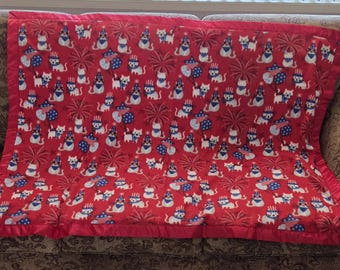 May include: Red blanket with a repeating pattern of white cats wearing patriotic hats and bow ties, with fireworks and starburst designs. The blanket has a red border and is placed on a patterned sofa.